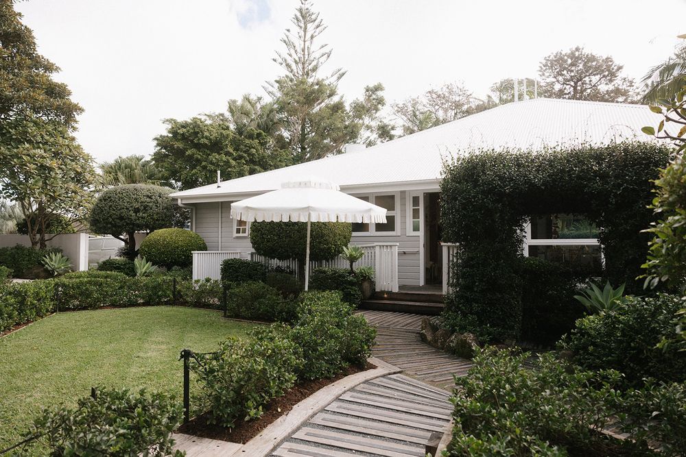 A modern white house exterior features a curved concrete walkway leading through a lush tropical garden landscape.