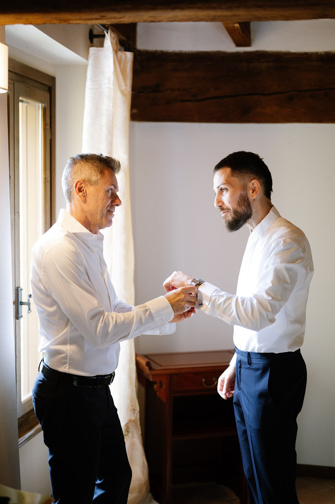 Two people in formal white shirts and blue pants stand in a sunlit room with wooden beams.