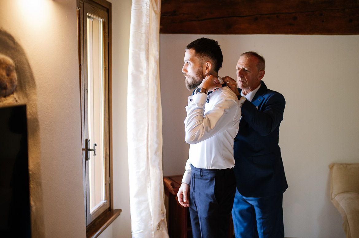 Two people getting ready in a sunlit room as one helps adjust the other's tie before a formal event.
