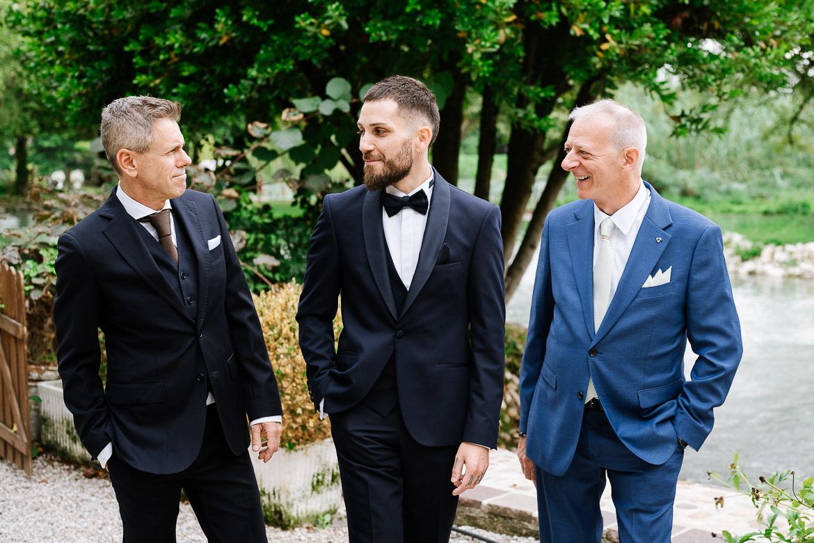 Groomsmen in formal attire walking and chatting along an outdoor path with greenery in the background.