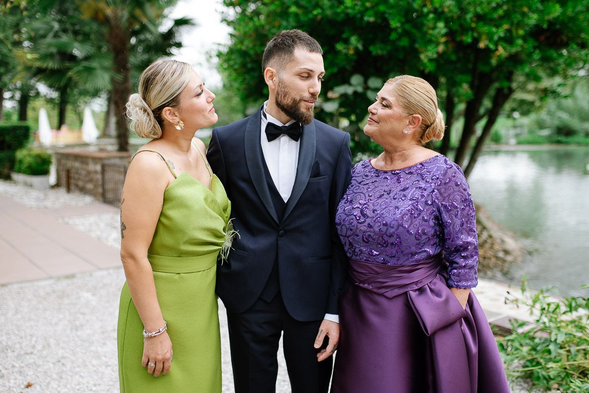 Family members dressed in formal attire gather outdoors near a pond for wedding photos.