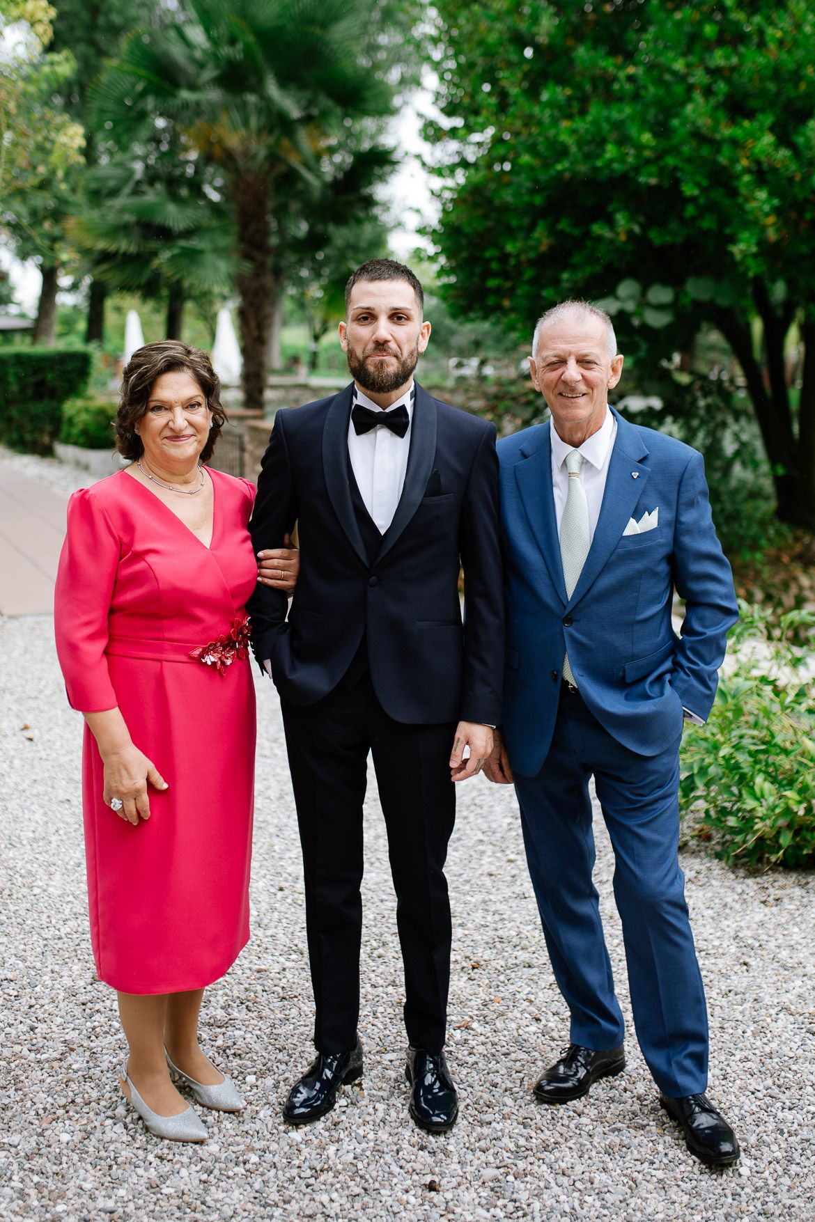 Group photo of wedding party members in formal attire standing on a gravel path surrounded by greenery.