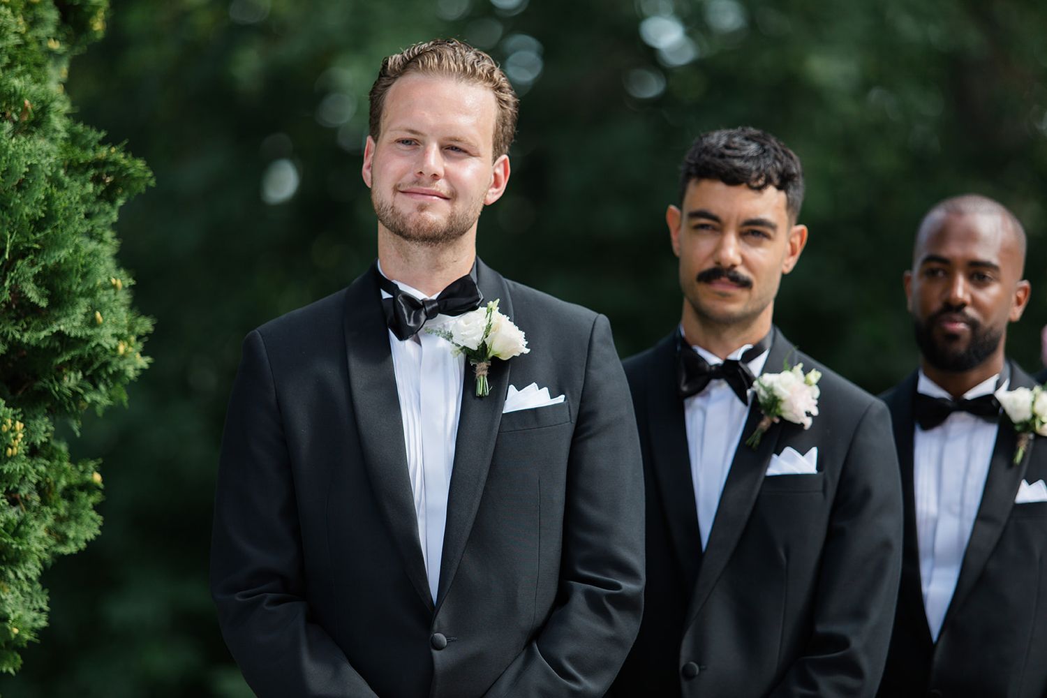 A row of groomsmen in black tuxedos and bowties stand together outdoors against greenery.