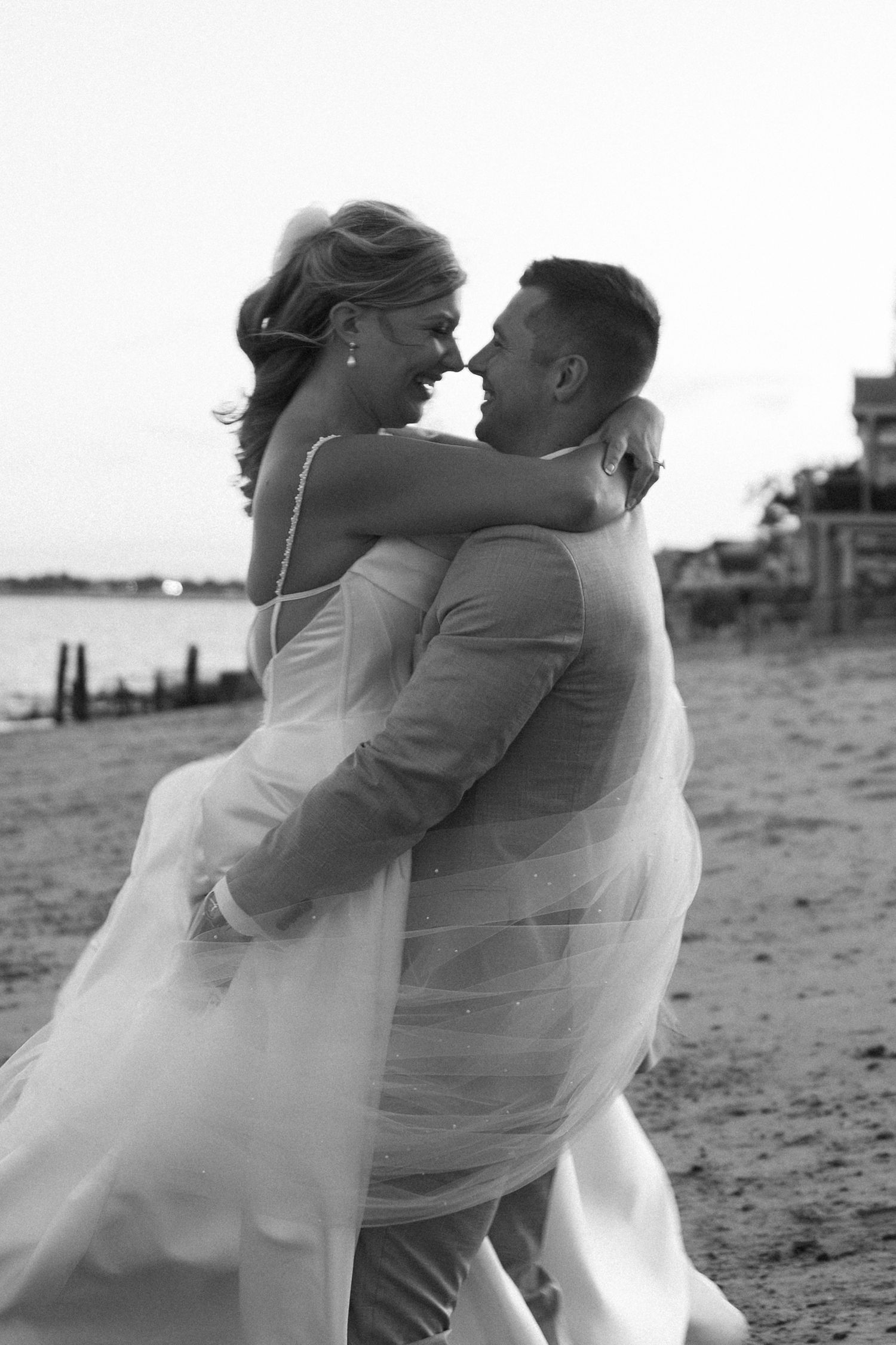 A romantic black and white beach wedding photo shows a couple embracing on the sand as her dress flows in the breeze.