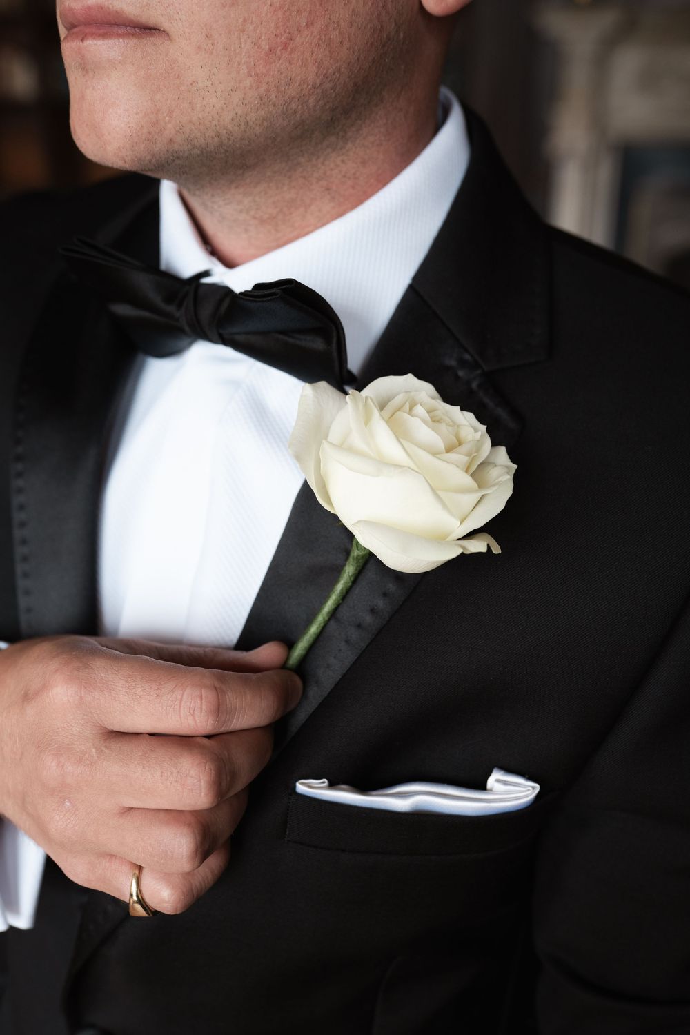 A white rose boutonniere is being pinned onto a black tuxedo lapel in an elegant wedding detail shot.