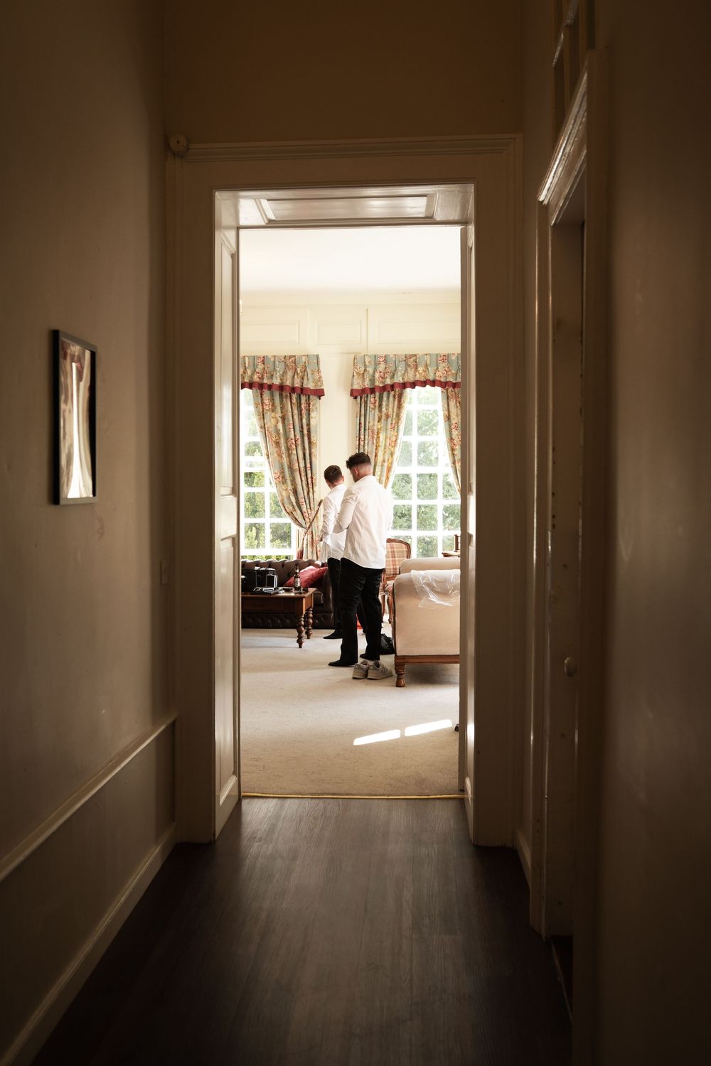 View through dark hallway into bright room with curtained windows and people standing by a window.