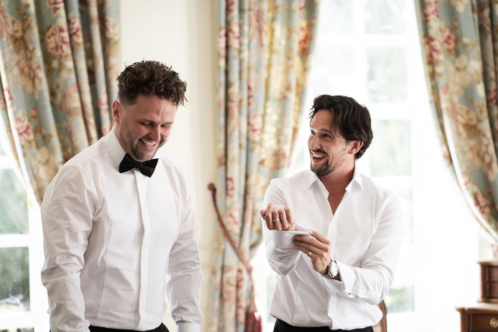 Two men in white dress shirts share a candid laughing moment together in an elegant room with floral curtains.