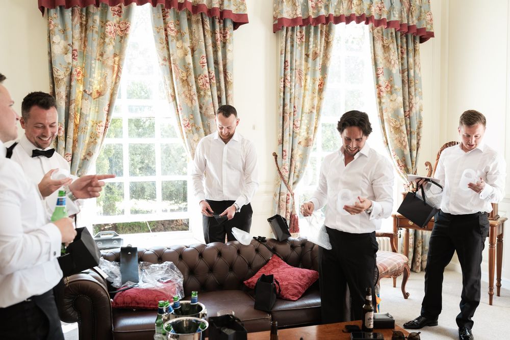 Groomsmen in white shirts prepare for a wedding ceremony in an elegant room with floral curtains.