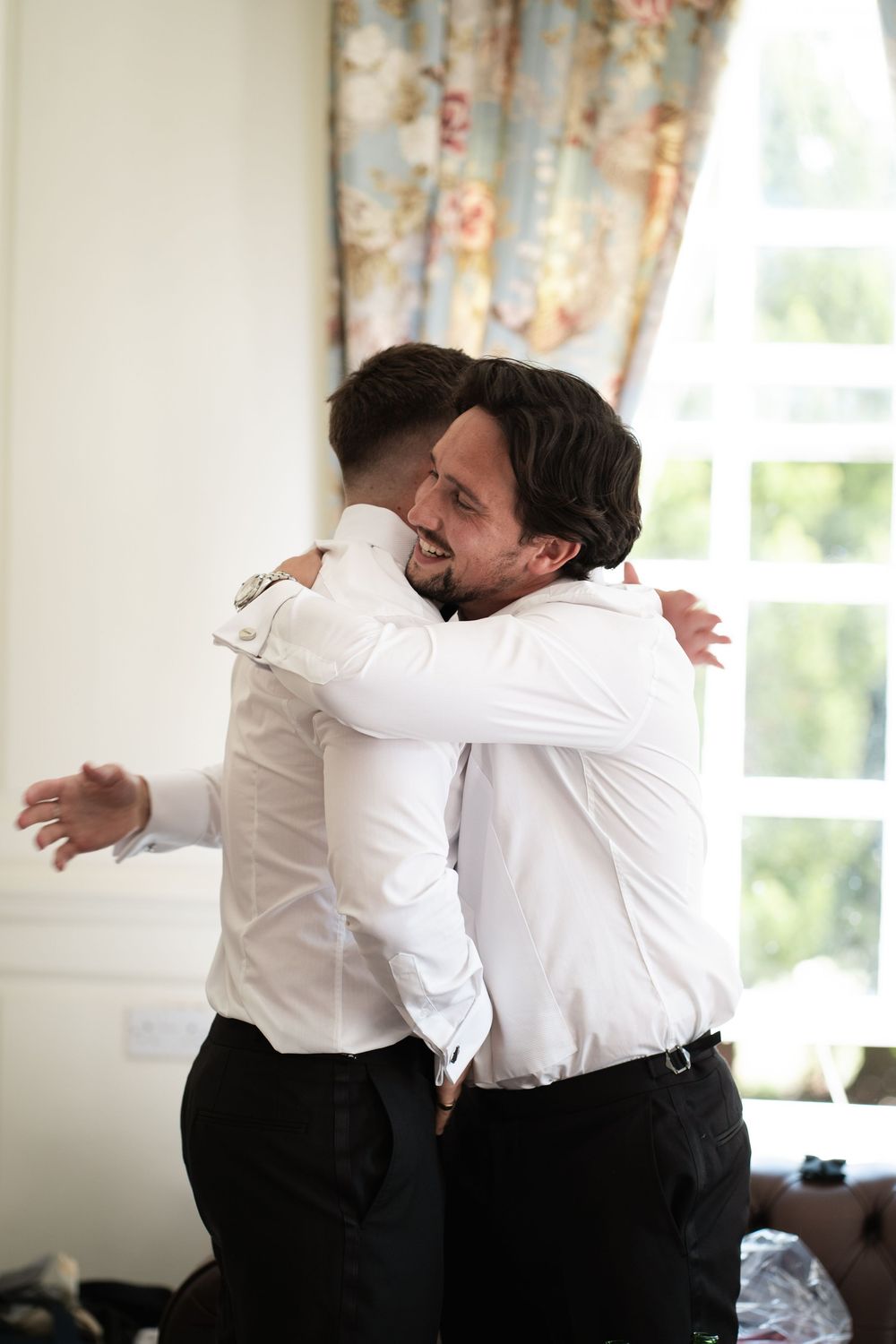Two people in white dress shirts and black pants embrace in a warm hug near a window.