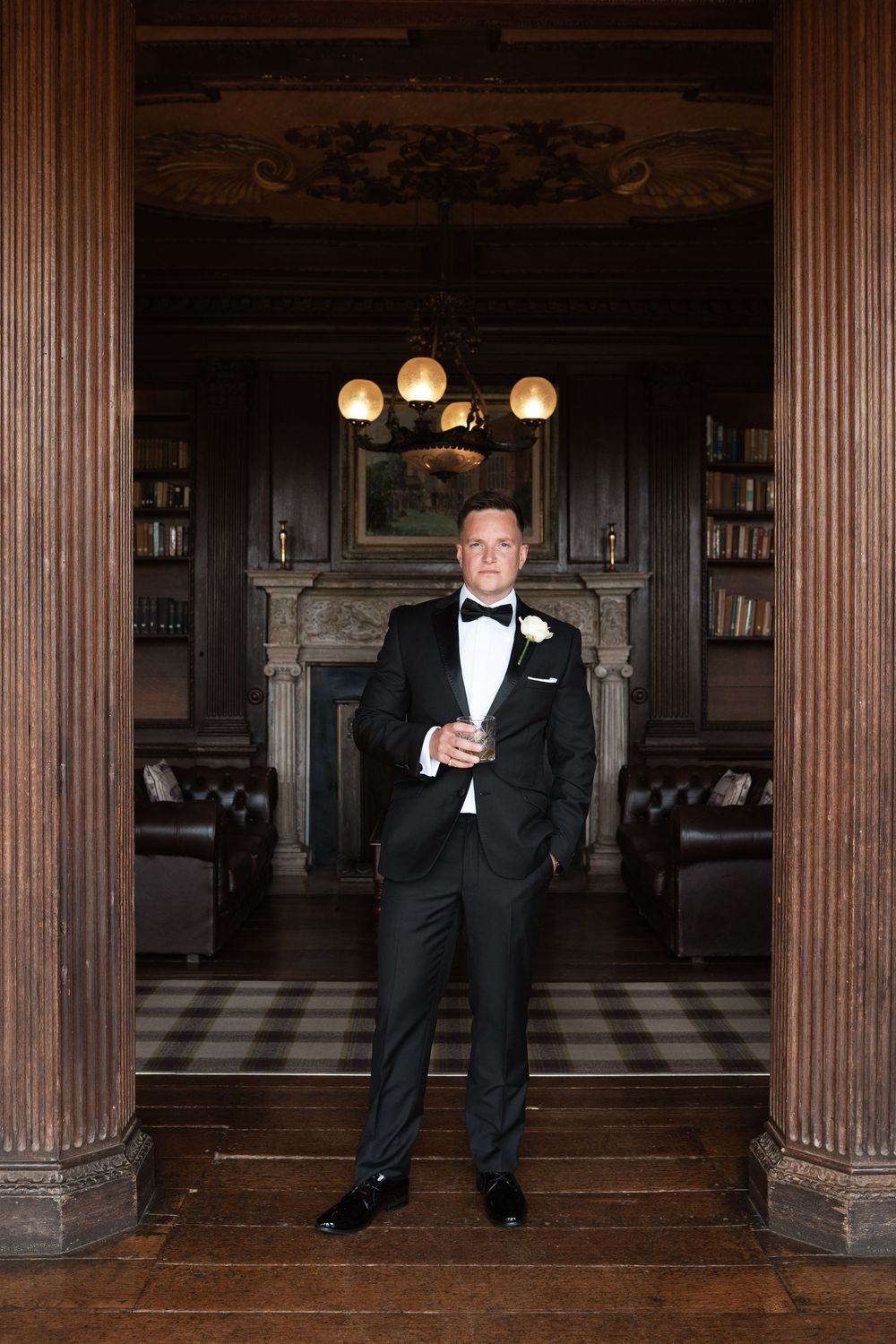 A groom in a classic black tuxedo poses between wooden columns in an elegant library setting.