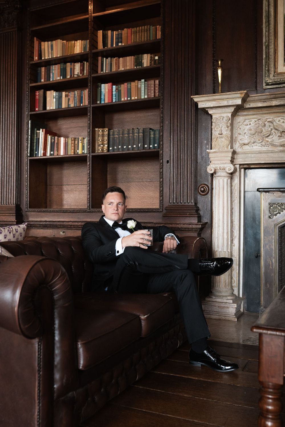 A person in a tuxedo relaxes on a leather couch in an elegant wood-paneled library with bookshelves.
