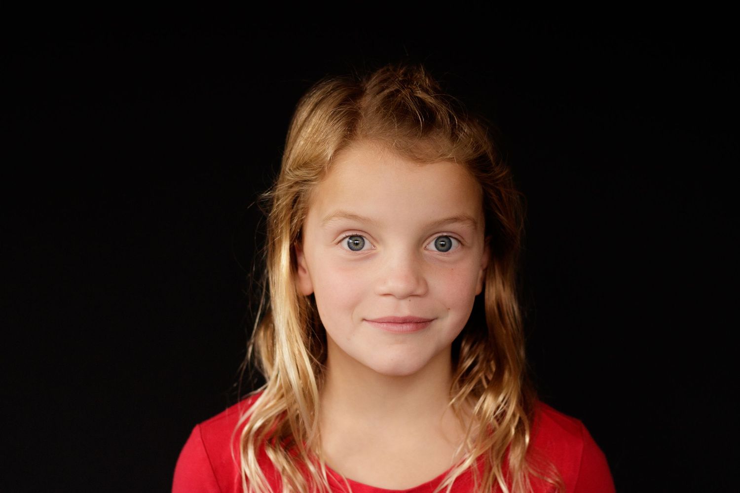 Young girl with long blonde hair and blue eyes smiling softly against a dark background.
