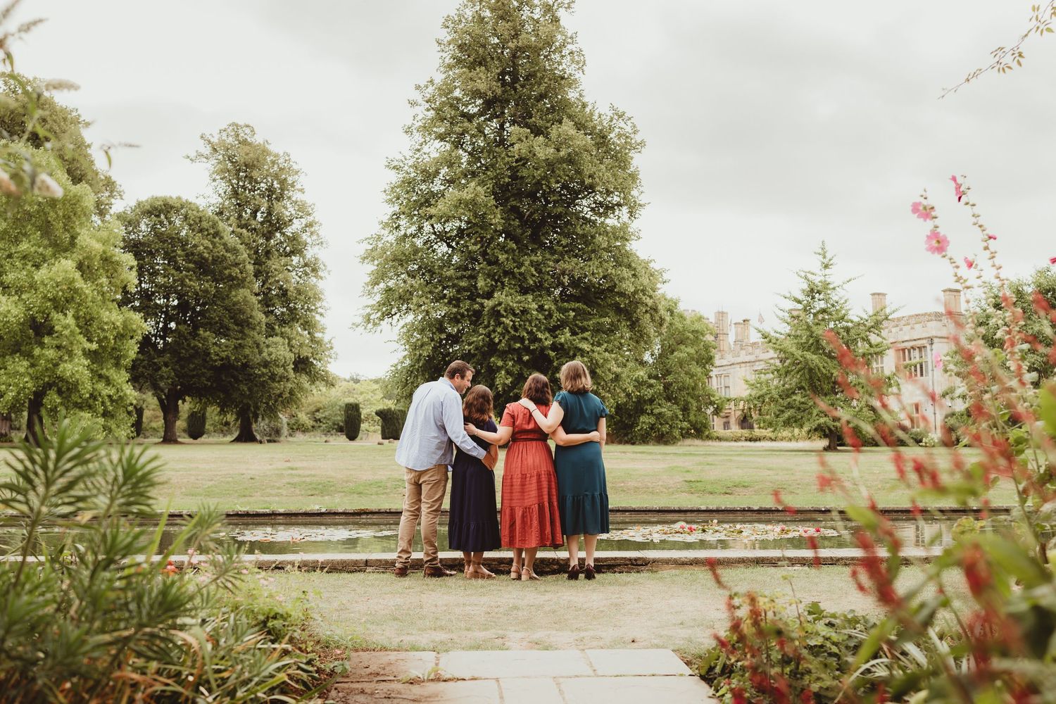 A group of people walk together on a grassy path under large oak trees near a historic stone building.