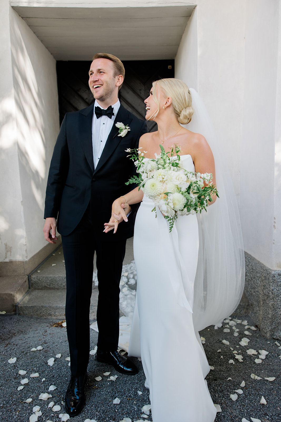 A joyful couple shares a candid moment in a white archway with scattered flower petals on the ground.
