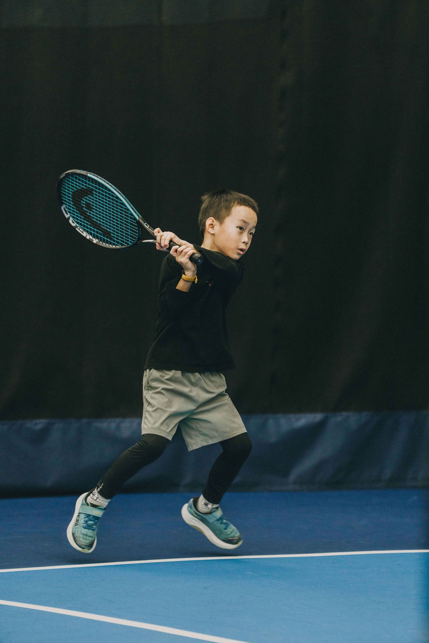 Young tennis player in grey shirt demonstrates proper serving form sequence on blue indoor court.