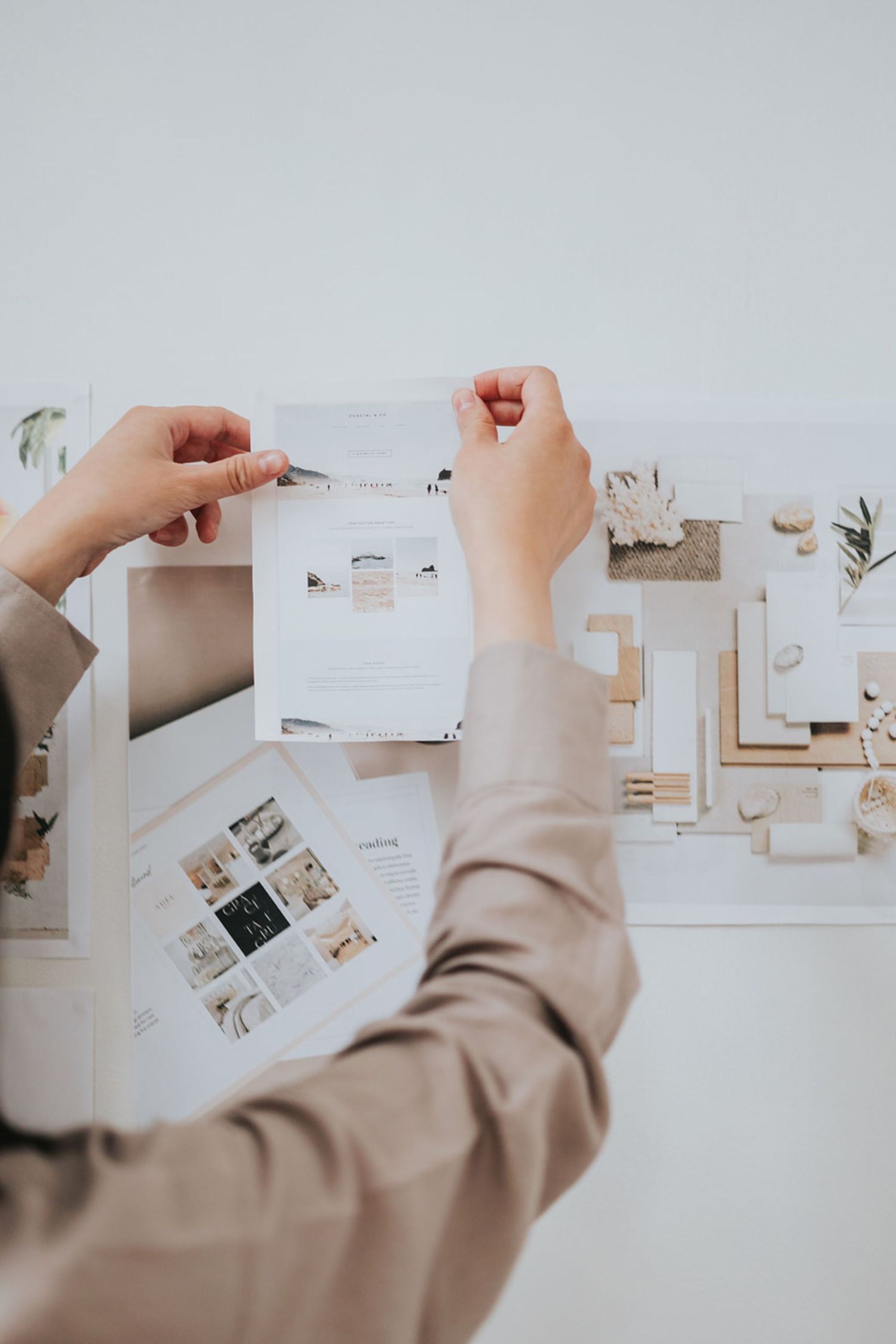 Hands arranging white paper cards and photos on a minimalist mood board against a light colored wall.