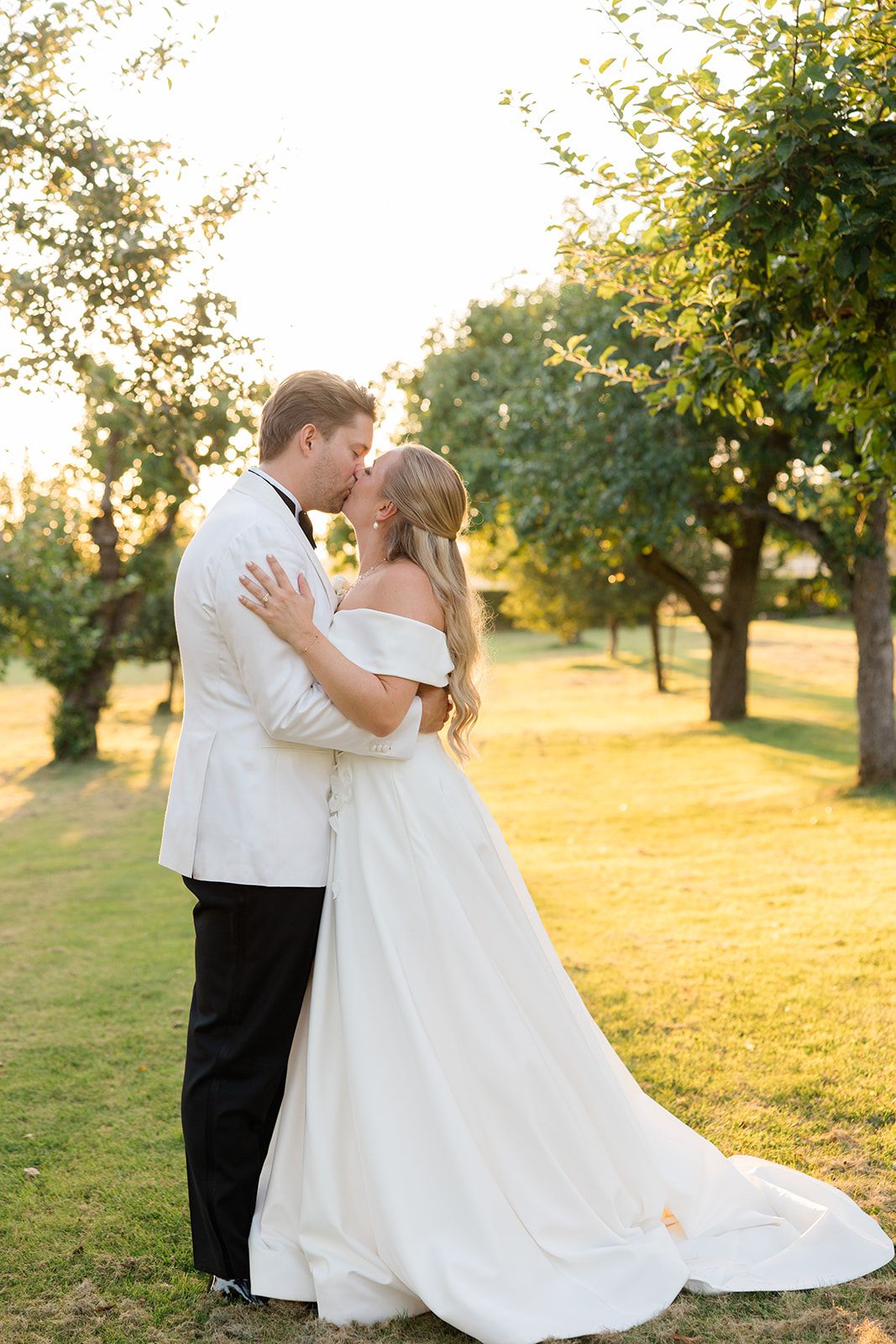 A couple in wedding attire embrace during golden hour in an orchard with sunlit trees in the background.