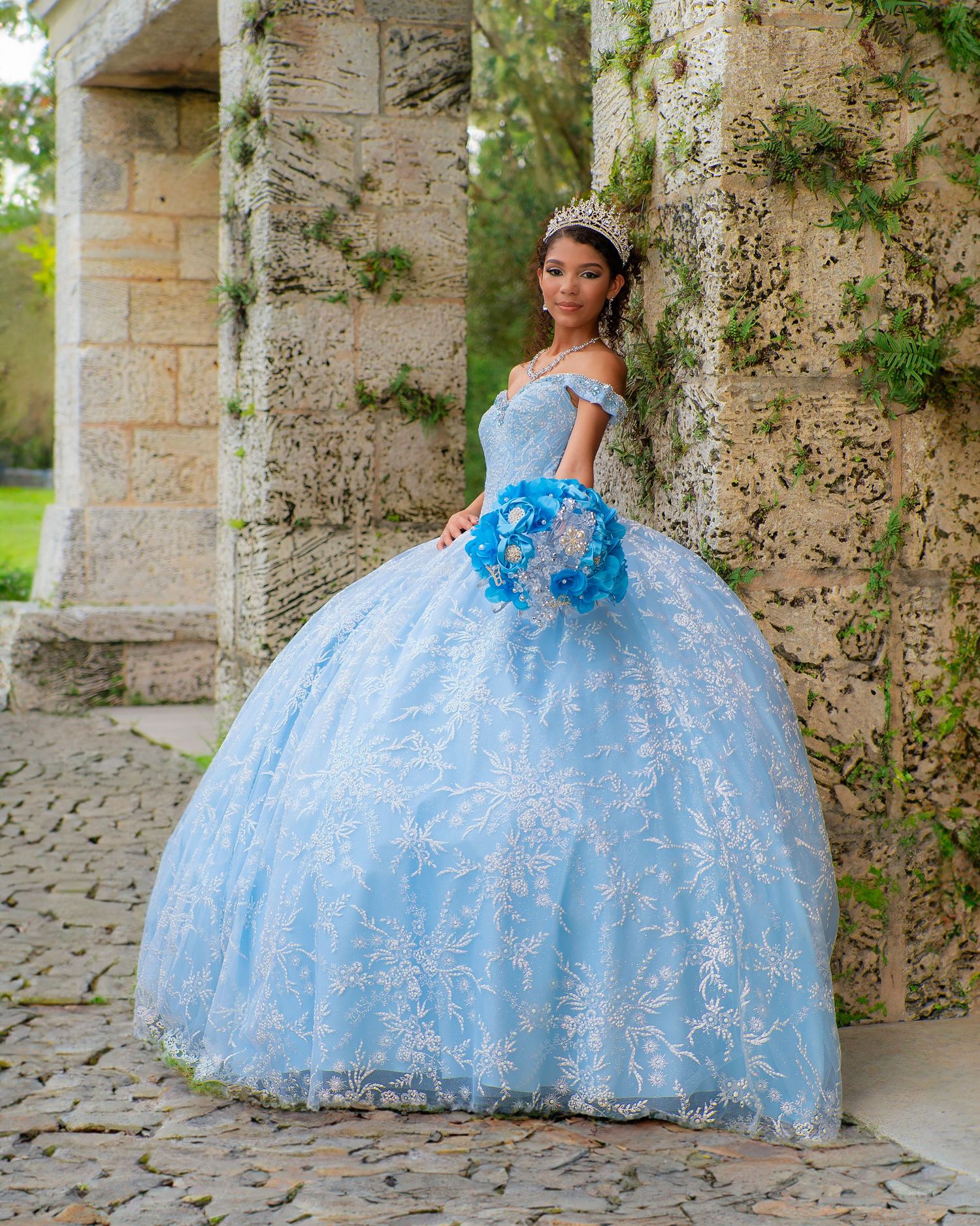Young woman in sky blue quinceañera ball gown holding blue bouquet beside stone columns outdoors.