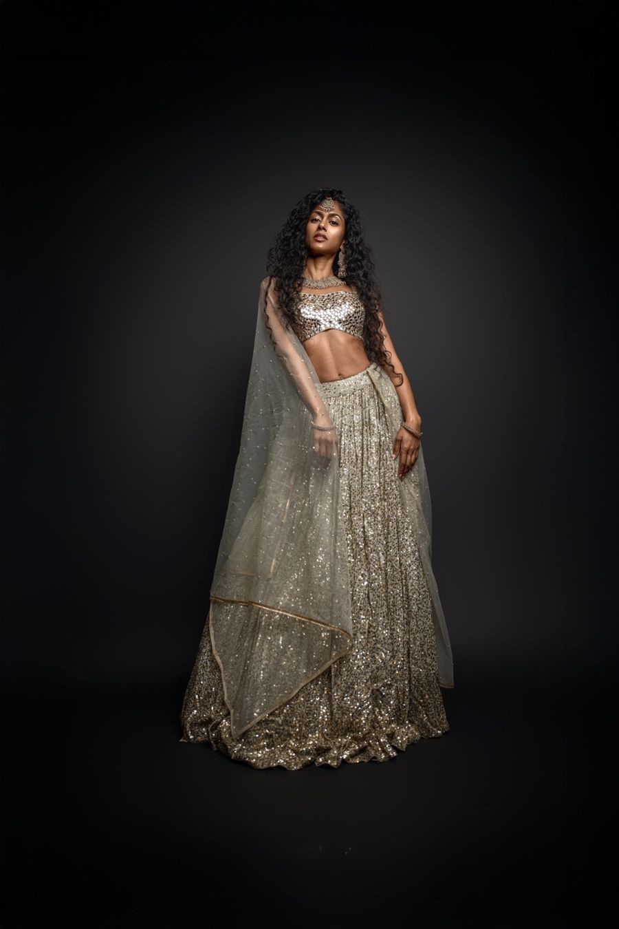 A person poses in a shimmering gold sequined dress with flowing fabric against a dark dramatic background.