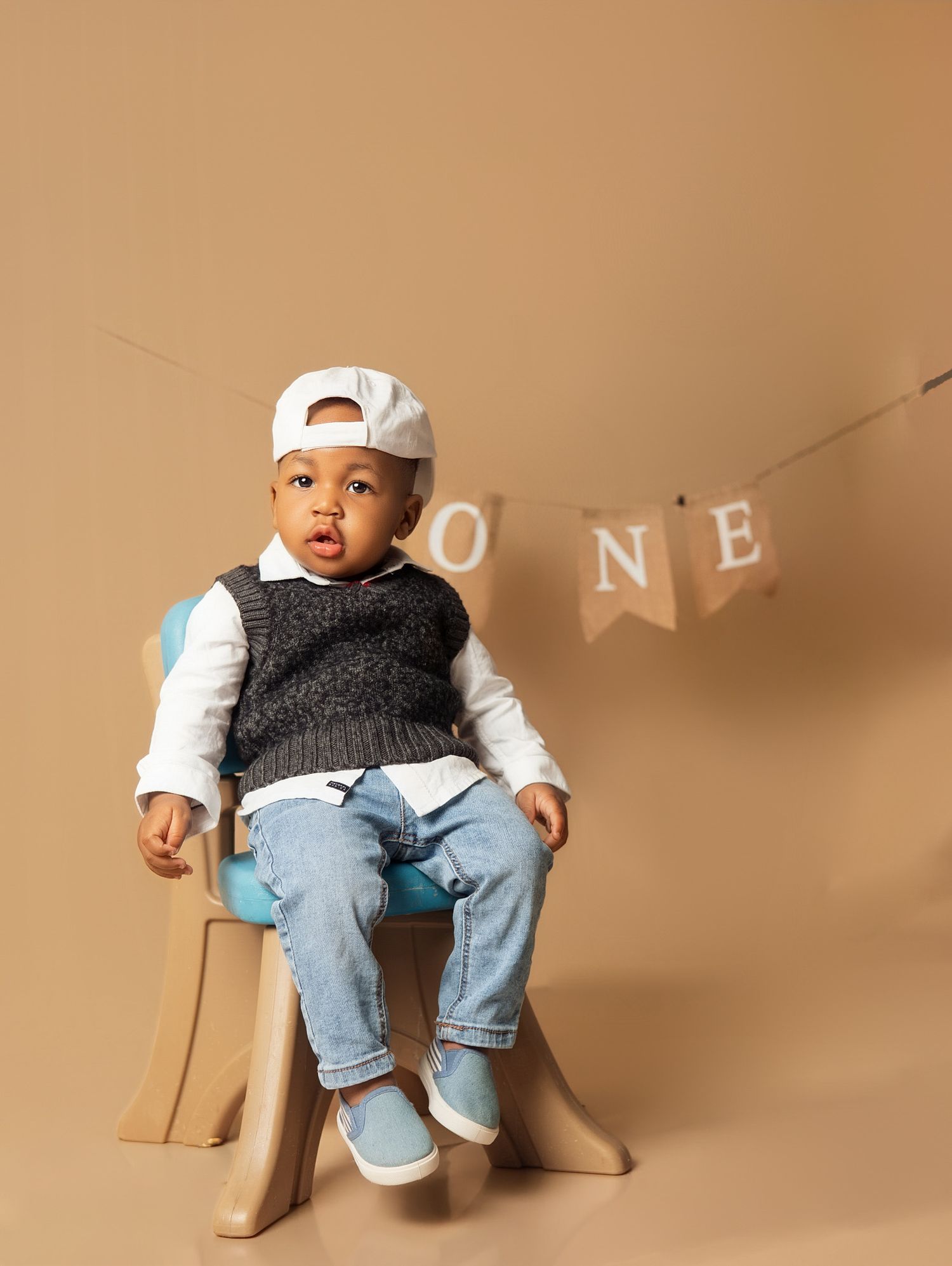 A baby boy in jeans, vest, and white cap sits on a small chair beside a ONE birthday banner on a beige background.