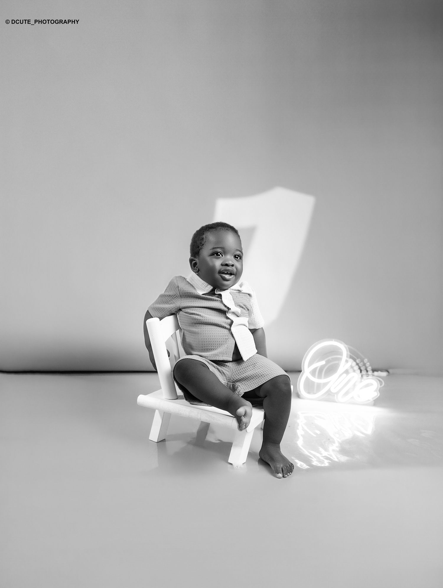 Black and white photo of a smiling toddler sitting on a small white chair beside a glowing number one neon sign.
