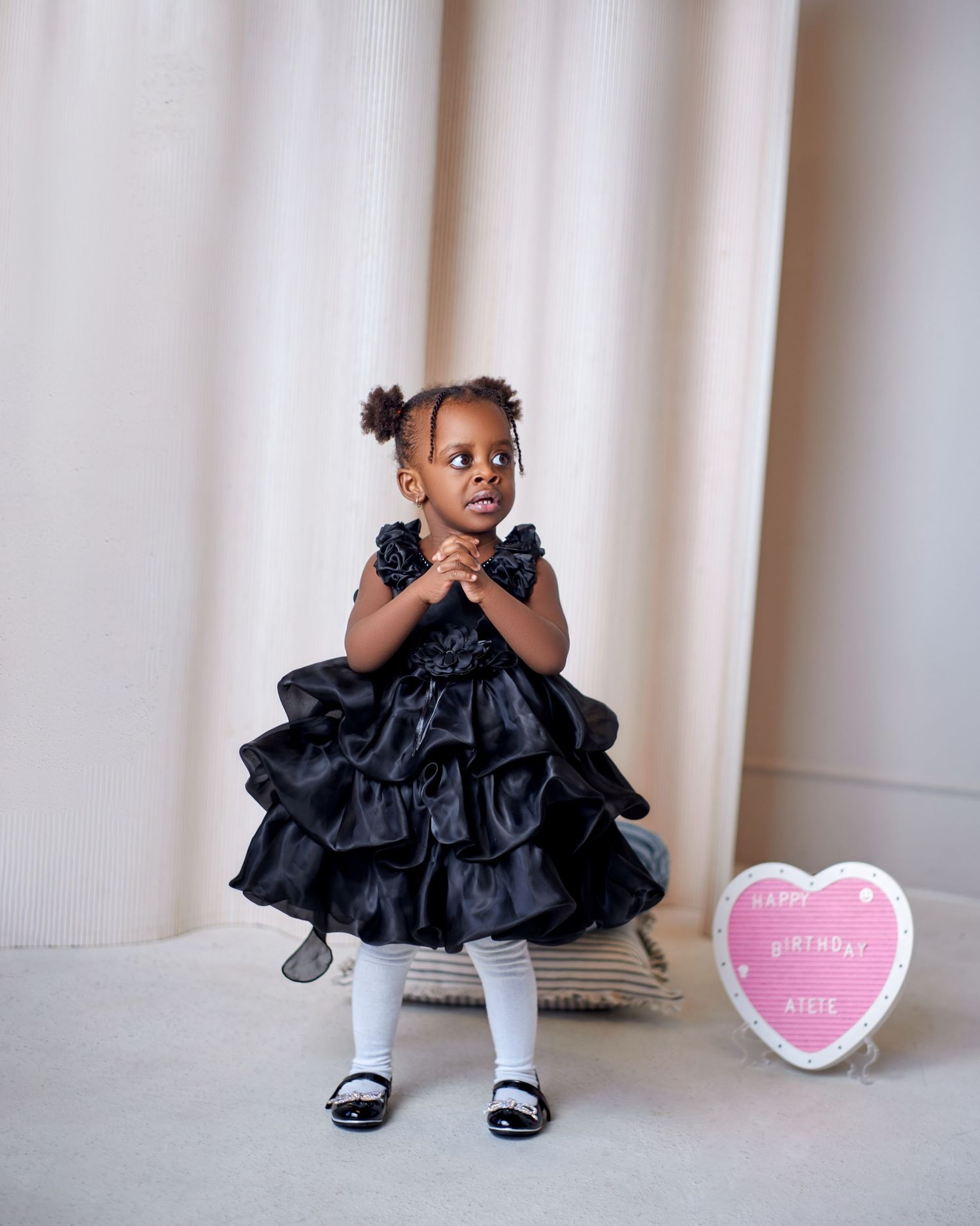 Adorable Black toddler girl in black ruffled dress stands beside a Happy Birthday heart sign.