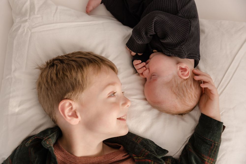 Two children resting peacefully together on white bedding in matching dark outfits.