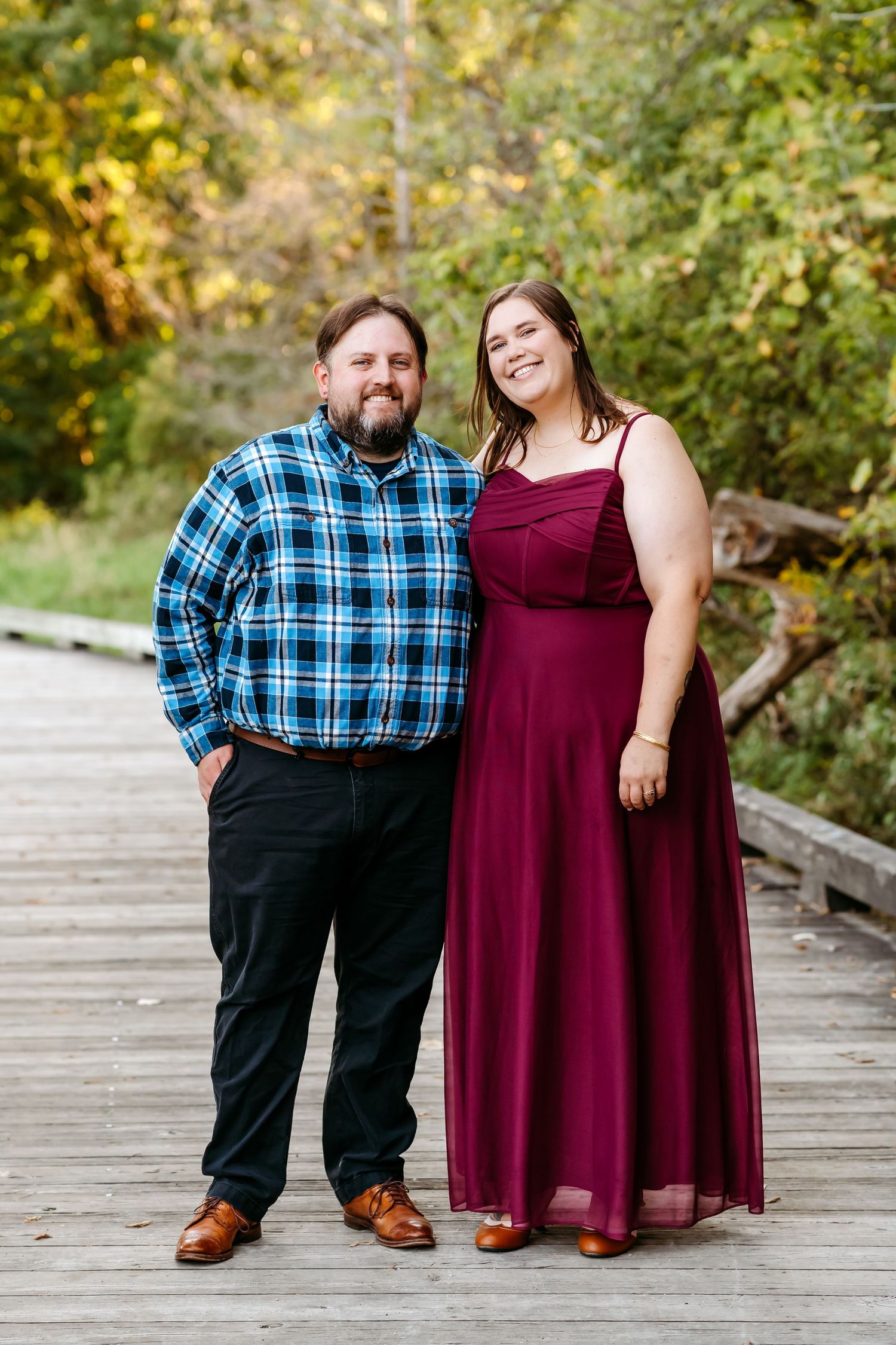 A couple in burgundy dress and plaid shirt pose together on a wooden boardwalk during an autumn photoshoot.
