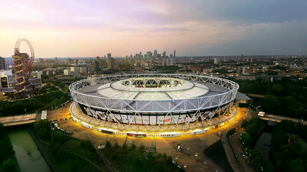 Aerial view of London Olympic Stadium and surrounding park area at dusk with glowing lights.