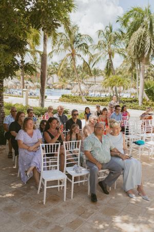 Wedding guests seated in white chairs under palm trees at an outdoor tropical venue.