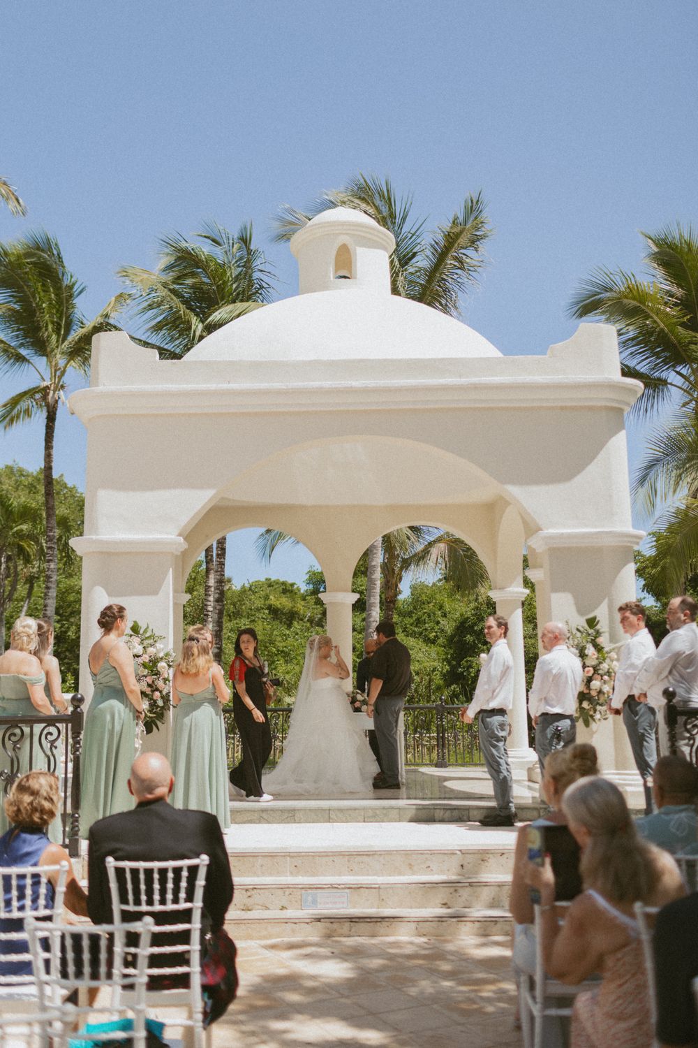 Wedding ceremony taking place under white Mediterranean arches with guests watching from white chairs.
