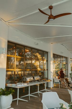 Modern restaurant interior with white decor, ceiling fans, and large windows overlooking tropical scenery.