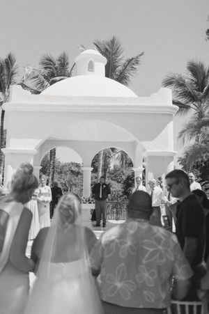 Wedding guests seated outdoors under white arches and palm trees at a tropical destination.