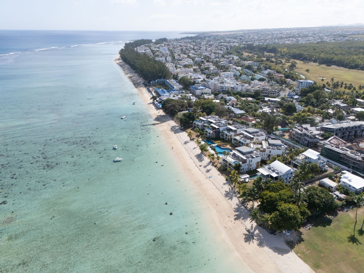 Aerial panoramic view of a tropical beach with turquoise waters, boats, and coastal buildings along the shoreline.