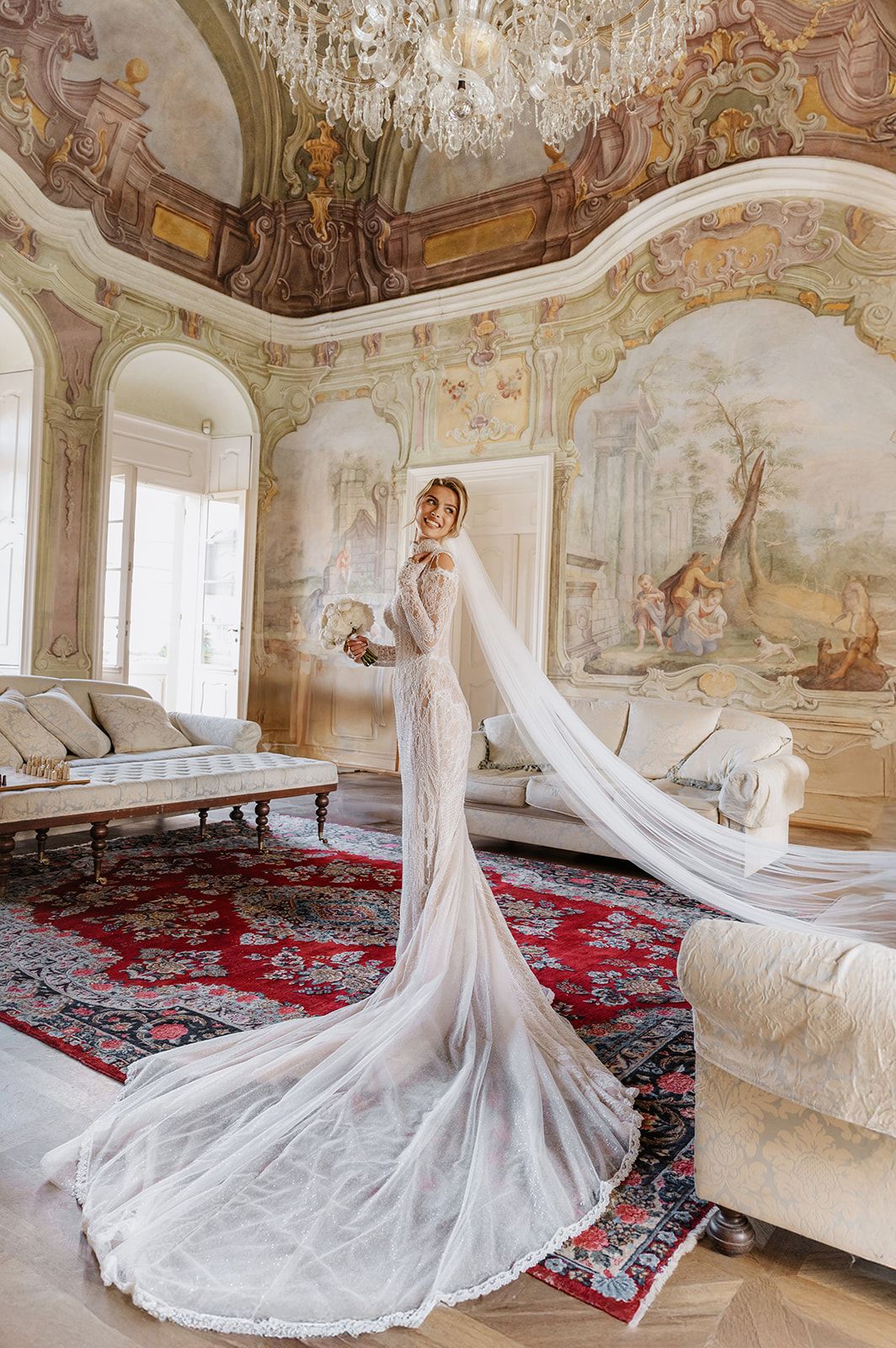 Wedding dress with dramatic long train displayed in ornate room with frescoed walls and crystal chandelier.