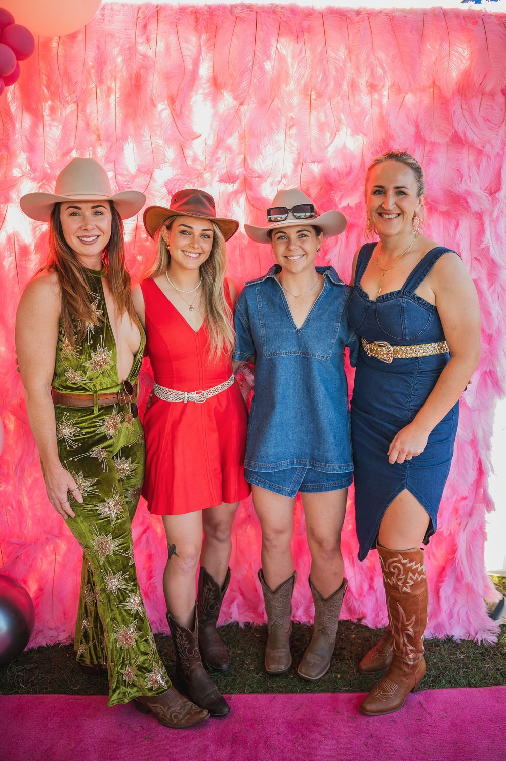 A group in cowboy boots and colorful dresses poses against a bright pink backdrop at a western-themed event.