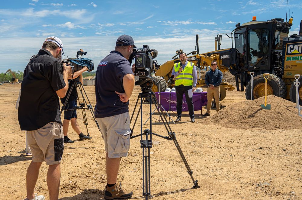 Camera operators film construction equipment and workers at a dusty outdoor site on a sunny day.