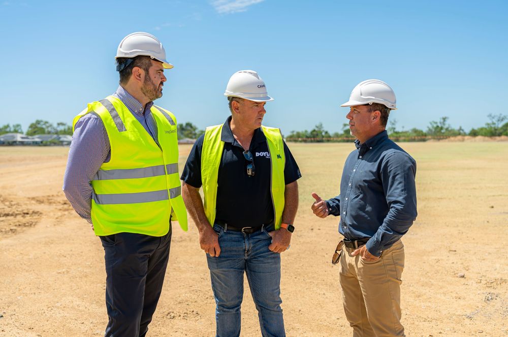 Construction workers in high visibility vests and hard hats discuss plans at an outdoor worksite on a sunny day.