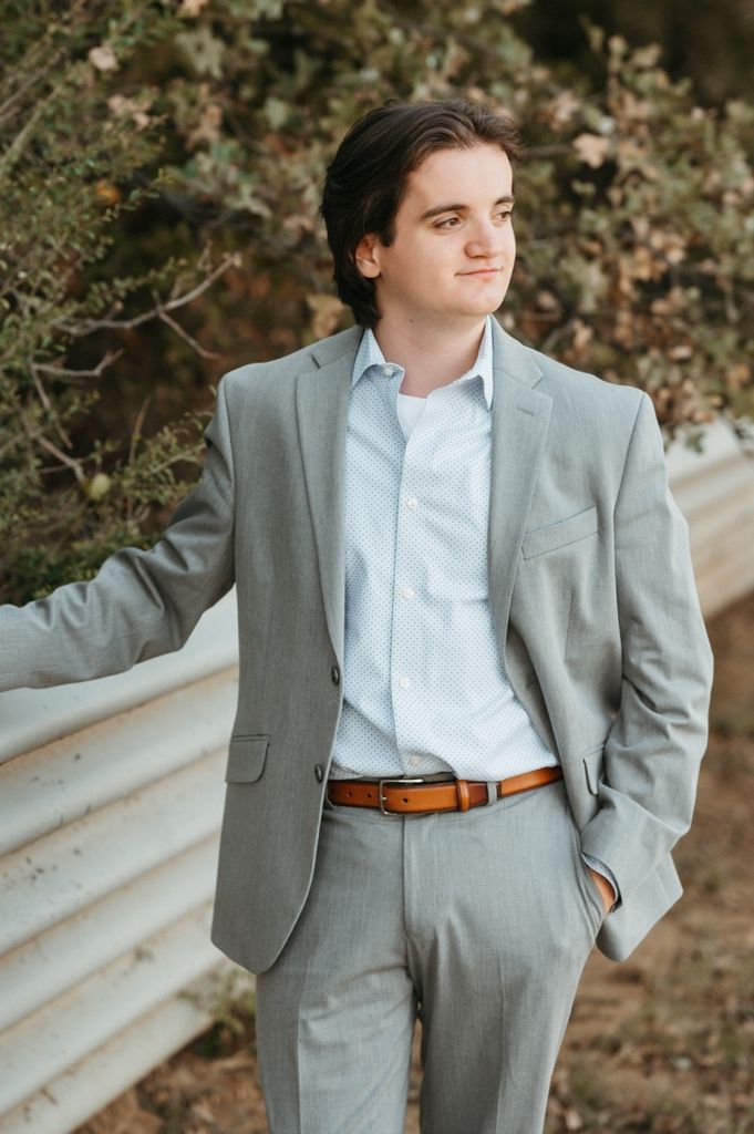 A person in a tailored gray suit and brown leather belt stands near a white railing outdoors.