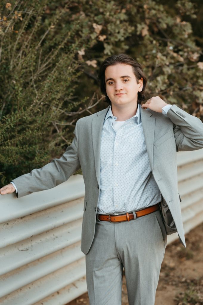 A person in a light gray suit and blue shirt poses against a blurred natural background in a professional photoshoot.