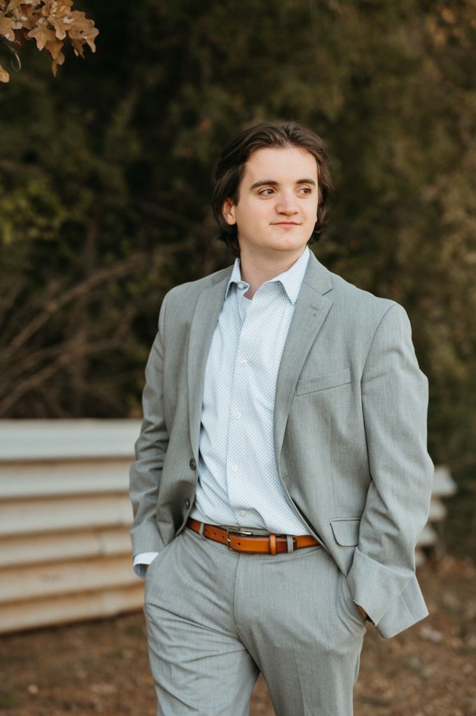 Professional portrait of someone in a light gray suit with brown leather belt and white dress shirt against autumn backdrop.
