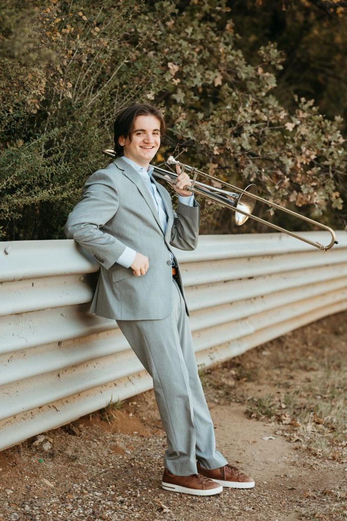 A musician in a light gray suit holds a trombone while standing outdoors against a rustic metal wall.