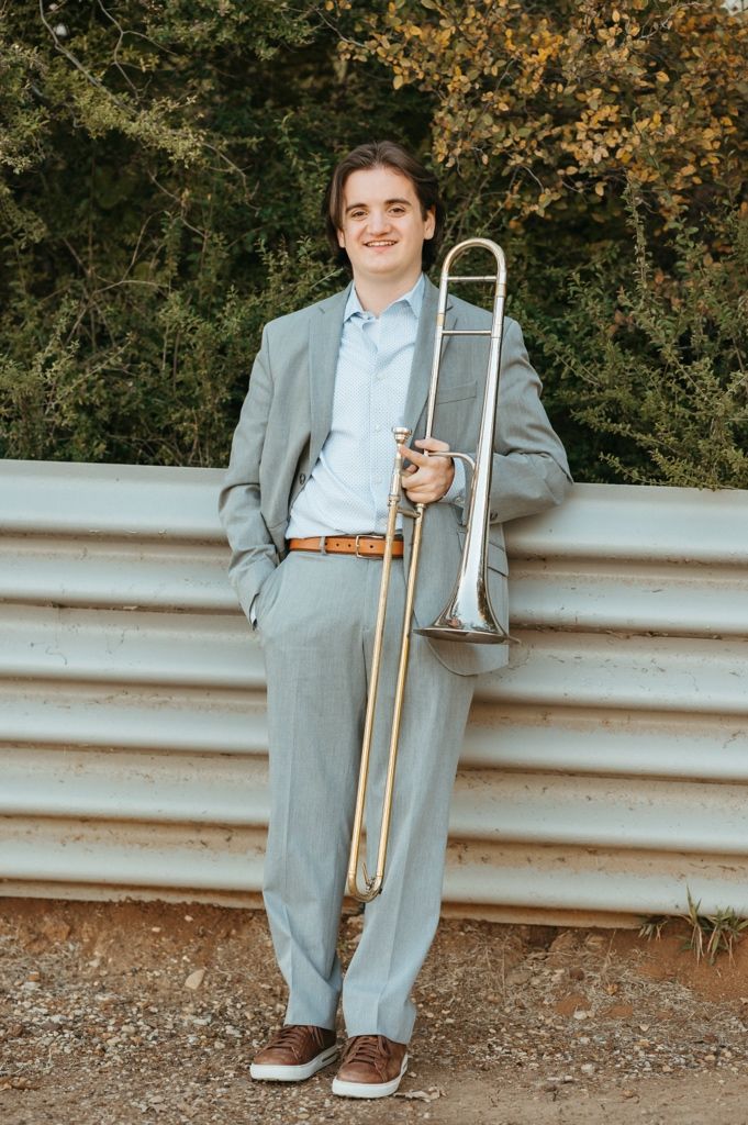 A musician in a gray suit poses with a trombone against a guardrail during an autumn outdoor photo session.