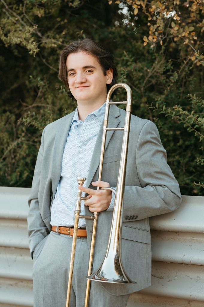 Musician in gray suit holding a trombone outdoors against autumn foliage background.
