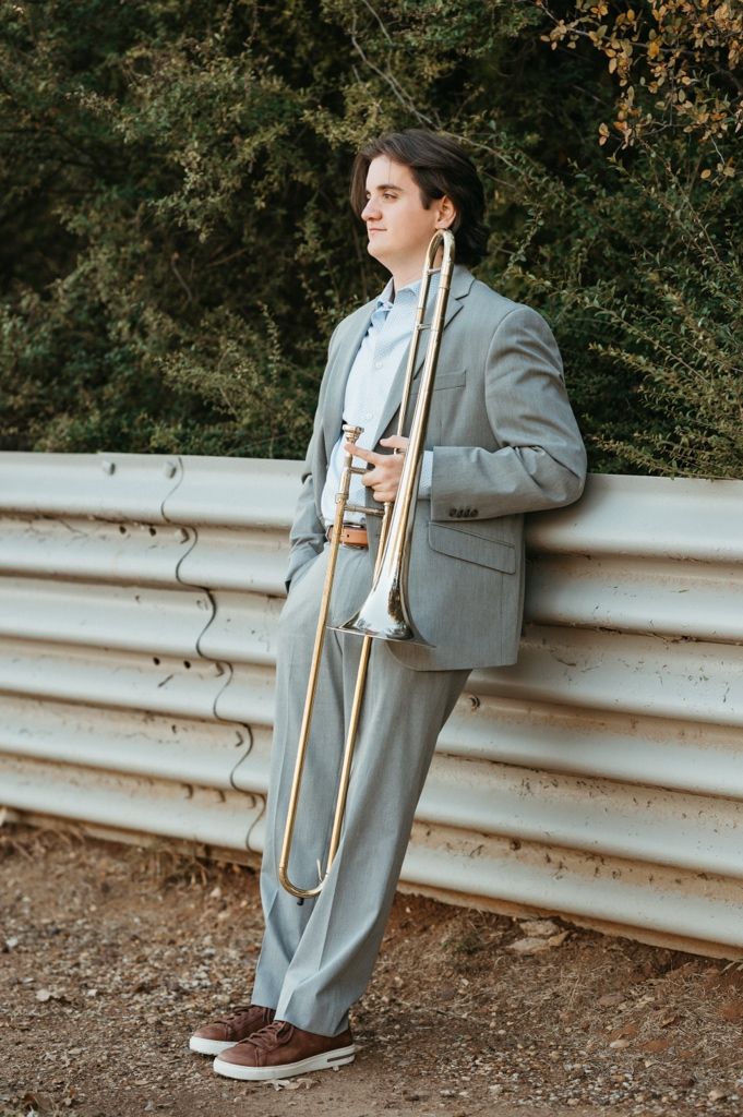 A musician in a gray suit leans against a guardrail while playing a trombone in an outdoor setting.