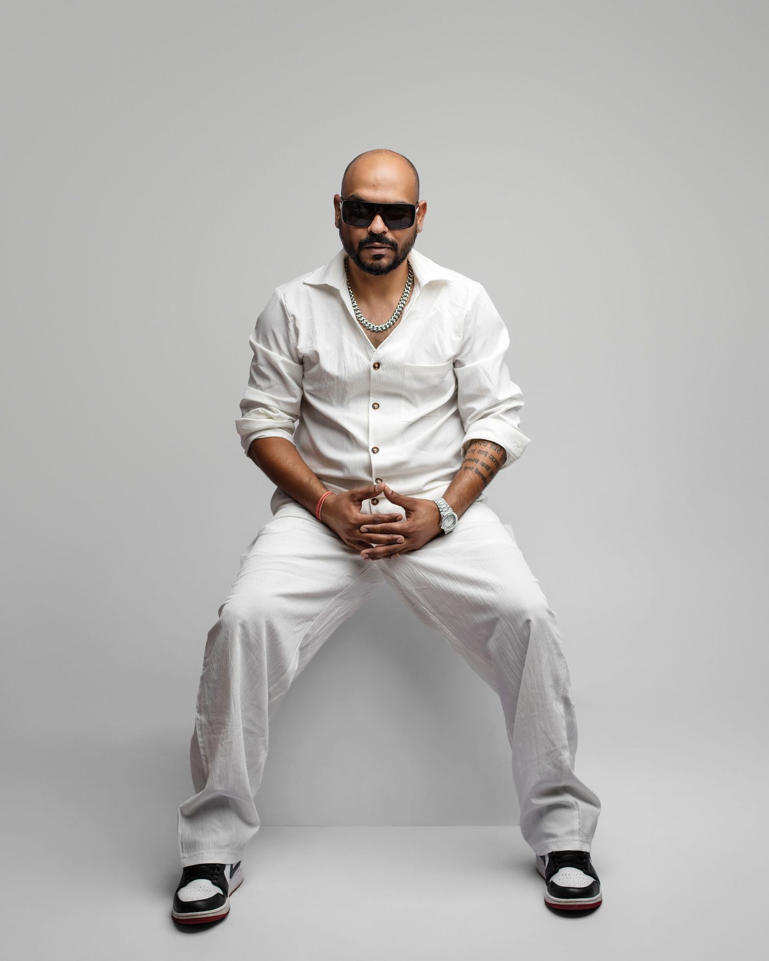 Figure in white clothing and sunglasses sits in casual pose on white background, creating minimalist aesthetic.