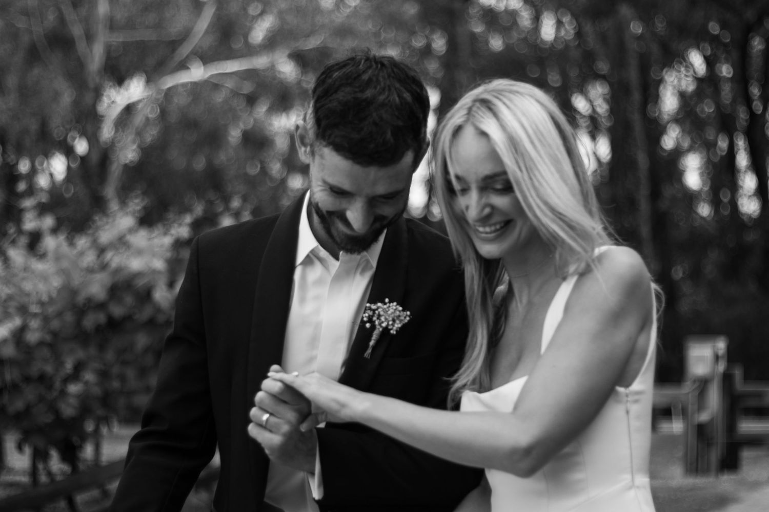 A black and white photo of a bride and groom smiling and holding hands as they walk outdoors together.