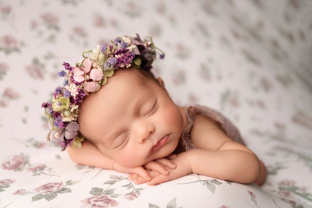 Sleeping newborn baby wearing purple floral bonnet resting on floral patterned blanket.