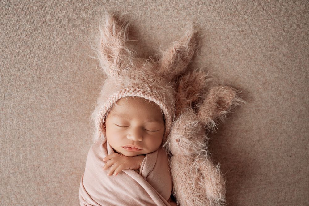 Sleeping newborn baby wearing a fuzzy pink bunny ear bonnet, wrapped in a soft pink swaddle on a beige backdrop.