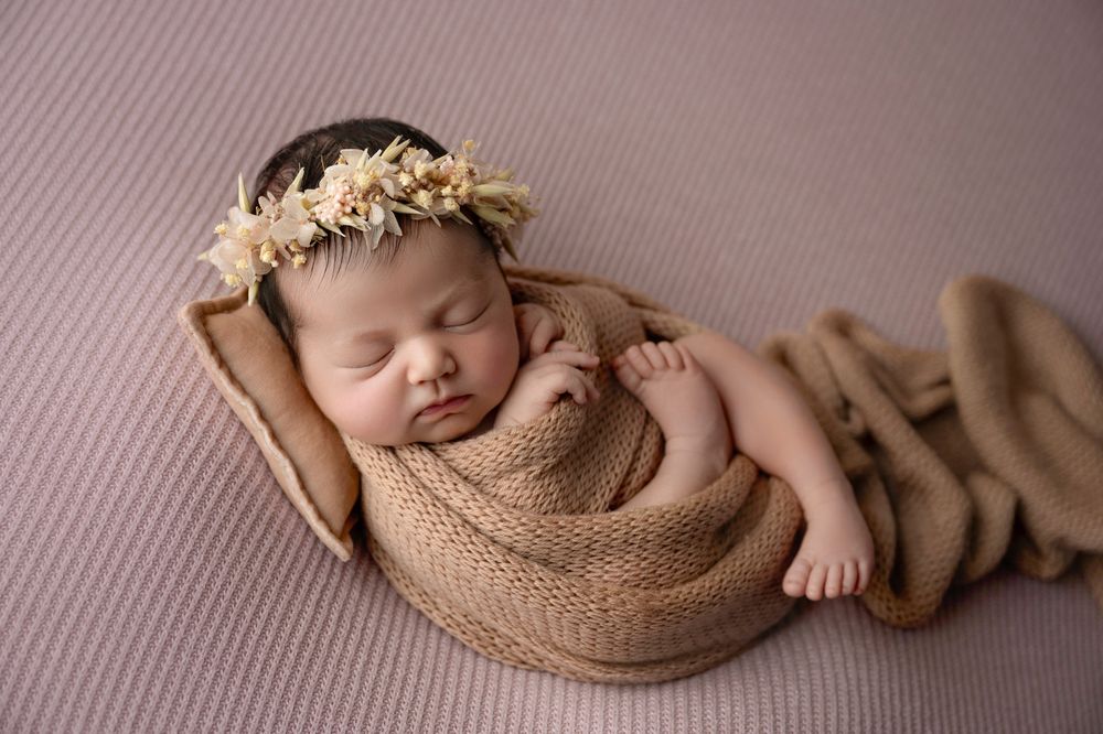 Newborn baby wrapped in tan knit blanket with floral headband sleeping in wooden dish.
